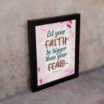 Christian wall art with brown 'Faith' and 'Fear' text, pink leaves, and a light pink floral background. Wall Art displayed on the wall in a black frame