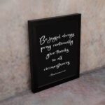 Bible verse wall art featuring 1 Thessalonians 5:16-18 in white cursive text on a solid black background, arranged clearly for easy reading. displayed on the wall in a black frame