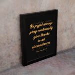 Bible verse wall art featuring 1 Thessalonians 5:16-18 in gold cursive text on a solid black background, arranged in a stacked format. displayed on the wall in a black frame