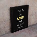 Psalm 37:3 Bible verse wall art with a black background, Lord highlighted in yellow. Christian home decor by Infidu. Wall Art kept on the wall in a black frame