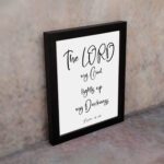 The Lord My God Lights Up My Darkness Psalm 18:28 white text on a black background with a black frame Christian wall art. displayed on the wall in a black frame