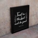 Trust In The Lord And Do Good Psalm 37:3 white text with decorative branches on black background Christian wall art displayed on the wall in black frame