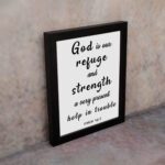 Psalm 46:1 Bible wall art with black bold text on a white background, saying 'God is our refuge and strength, a very present help in trouble. Wall Art displayed on the wall in a black frame