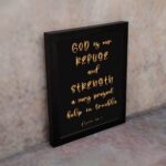 Psalm 46:1 Bible wall art with gold handwritten text on a black background, saying 'God is our refuge and strength. Wall Art displayed on the wall in a black frame