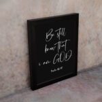 Be Still and Know That I Am God - Psalm 46:10 Bible Verse Wall Art with White Cursive Text on Black Background Wall Art displayed on the wall in a black frame