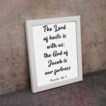 The Lord Of Hosts Is With Us - Psalm 46:7 Bible Verse Wall Art with White Text on Light Background Framed in Black Wall Art displayed on the wall in a white frame