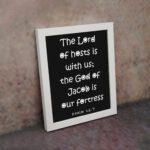 The Lord Of Hosts Is With Us - Psalm 46:7 Bible Verse Wall Art with White Text on Black Background Wall Art displayed on the wall in a white frame