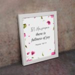 Psalm 16:11 wall art with a white background and pink flower petals, featuring the text 'In His presence there is fullness of joy. Wall Art displayed on the wall in a white frame