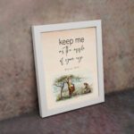 Psalm 17:8 wall art with a soft cursive font and a peaceful illustration of a person sitting under a tree. Wall Art is displayed on the wall in a white frame.