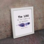 The Lord My God Lights Up My Darkness Psalm 18:28 Bible Verse Wall Art with black text and a purple and blue feather on a white background. Wall Art displayed on the wall in a white frame