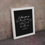 Black wall art with white text and small flower designs quoting When you go through deep waters I will be with you from Isaiah 43:2. displayed on the wall in a white frame