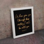 Black wall art with gold cursive text reading When you go through deep waters I will be with you from Isaiah 43:2. displayed on the wall in a white frame