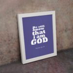 Be Still and Know That I Am God Psalm 46:10 Christian Wall Art with bold white text on a dark purple background. Wall Art displayed on the wall in a white frame