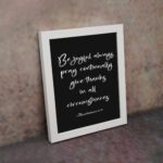 Bible verse wall art featuring 1 Thessalonians 5:16-18 in white cursive text on a solid black background, arranged clearly for easy reading. displayed on the wall in a white frame