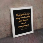 Bible verse wall art featuring 1 Thessalonians 5:16-18 in gold cursive text on a solid black background, arranged in a stacked format. displayed on the wall in a white frame