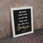 Psalm 63:3 Bible verse wall art with a dark background and cursive Glorify You in light gold. Christian home decor by Infidu. Wall Art kept on the wall in a white frame