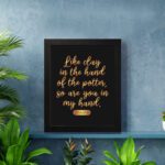 Infidu wall art with the text Like clay in the hand of the potter, so are you in my hand in golden letters on a black background. displayed on the wall in a black frame