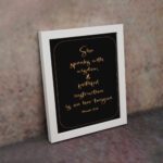 Proverbs 31:26 Bible wall art with gold text on a black background, framed by a golden border. Featuring elegant cursive font. displayed on the wall in a white frame