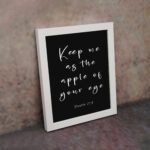 Keep Me As The Apple Of Your Eye Psalm 17:8 white cursive text on black background - Christian wall art for home decor displayed on the wall in a white frame