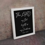 The Lord My God Lights Up My Darkness Psalm 18:28 white handwritten text on black background Christian wall art displayed on the wall in a white frame