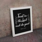 Trust In The Lord And Do Good Psalm 37:3 white text with decorative branches on black background Christian wall art displayed on the wall in white frame