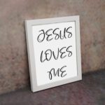 Jesus Loves Me Bible wall art with black script text on a white background. Wall Art displayed on the wall in a white frame