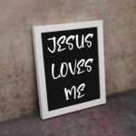 Jesus Loves Me Bible wall art with bold gold text and a decorative gold border on a black background. Wall Art displayed on the wall in a white frame