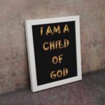 I Am A Child Of God Bible wall art with bold golden yellow text on a black background. Wall Art displayed on the wall in a white frame