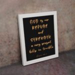 Psalm 46:1 Bible wall art with gold handwritten text on a black background, saying 'God is our refuge and strength. Wall Art displayed on the wall in a white frame