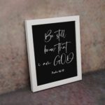 Be Still and Know That I Am God - Psalm 46:10 Bible Verse Wall Art with White Cursive Text on Black Background Wall Art displayed on the wall in a white frame