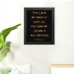 The Lord Of Hosts Is With Us - Psalm 46:7 Bible Verse Wall Art with Golden Text on Black Background Wall Art hung on the wall in a black frame