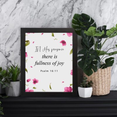 Psalm 16:11 wall art with a white background and pink flower petals, featuring the text 'In His presence there is fullness of joy. Wall Art displayed on the table in a black frame