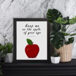 Keep Me As The Apple Of Your Eye" Psalm 17:8 Bible Wall Art featuring a red apple and white background. Perfect Christian home decor. Wall Art displayed on the table in black frame