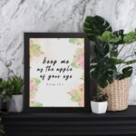 Keep Me As The Apple Of Your Eye Psalm 17:8 Bible Verse Wall Art with apple and leaf design, soft pastel colors, and white background. Wall Art displayed on the table in black frame