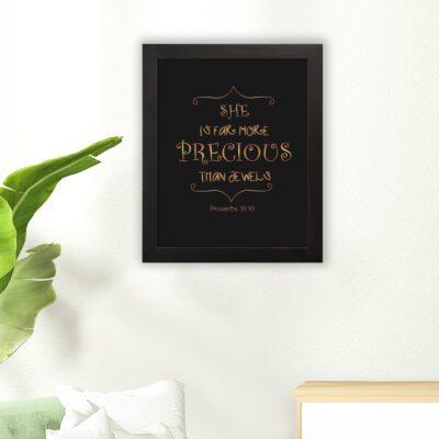 Black wall art with gold text saying SHE IS FAR MORE PRECIOUS THAN JEWELS from Proverbs 31:10, styled with decorative elements. hung on the floor in a black frame