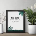 The Lord My God Lights Up My Darkness Psalm 18:28 Bible Verse Wall Art with black text and green tropical leaves on a white background. Wall Art displayed on the table in a black frame