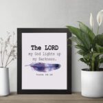 The Lord My God Lights Up My Darkness Psalm 18:28 Bible Verse Wall Art with black text and a purple and blue feather on a white background. Wall Art displayed on the table in a black frame