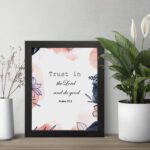 Trust In The Lord And Do Good Psalm 37:3 Bible Verse Wall Art with watercolor and floral designs on a white background. Wall Art displayed on the wall in a black frame