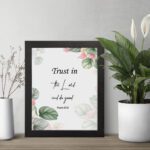 Trust in the Lord and Do Good Psalm 37:3 Bible Wall Art with botanical green leaves and pink flowers on a white background. Wall Art displayed on the table in a black frame