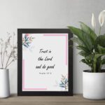 Infidu Bible wall art featuring Psalm 37:3 in black text with blue and pink floral accents and a soft pink border. Wall Art displayed on the table in a black frame