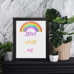 Infidu Jesus Loves Me Bible Wall Art with rainbow arc and colorful text in purple, yellow, and pink fonts Wall Art displayed on the table in a white frame