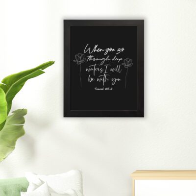 Black wall art with white text and small flower designs quoting When you go through deep waters I will be with you from Isaiah 43:2. hung on the floor in a black frame