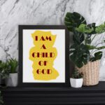 Infidu I Am A Child Of God Bible Wall Art with yellow shape and red text on a white background Wall Art displayed on the table in a black frame