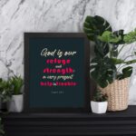 Infidu Psalm 46:1 Bible Wall Art with a deep blue background, pink and white text, featuring God is our refuge and strength. Wall Art displayed on the table in a black frame