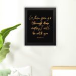 Black wall art with gold cursive text reading When you go through deep waters, I will be with you from Isaiah 43:2. hung on the floor in a black frame