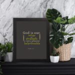 Psalm 46:1 Bible Wall Art with muted text on a dark gray background, featuring varying fonts and sizes. Wall Art displayed on the table in a black frame