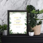 Psalm 46:1 Bible Wall Art with gold and black text, leafy green borders, and a white background. Wall Art displayed on the table in a black frame