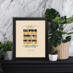 Psalm 50:15 Bible Wall Art with bold black, orange, and yellow text and simple floral embellishments on a cream background. Wall Art displayed on the table in a black frame