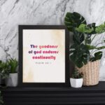 The Goodness Of God Endures Continually Psalm 52:1 Christian Wall Art with bold black and red text on a light cream background. Wall Art displayed on the table in a black frame