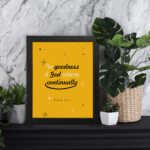 Bright mustard yellow wall art with Psalm 52:1. God in gold and playful font with star-like designs. Wall Art displayed on the table in a black frame